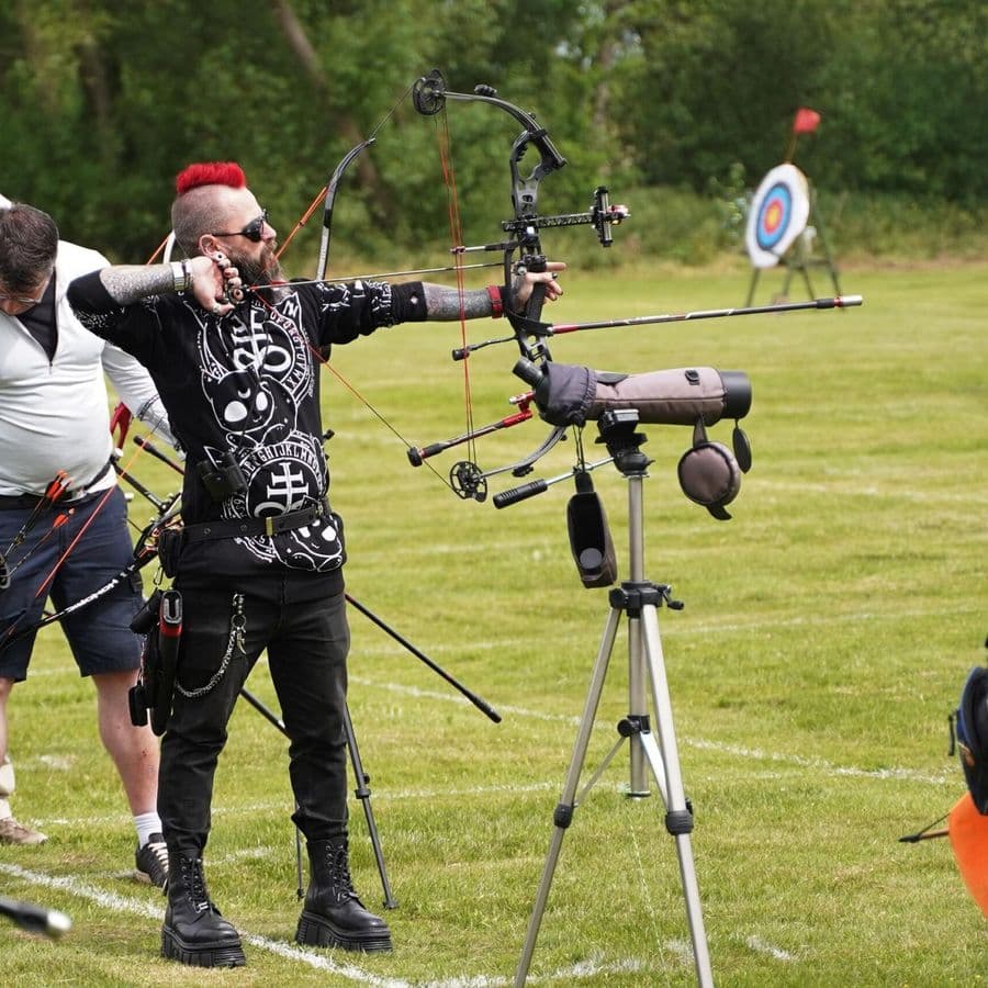An archer with a red mohawk and tattoos at full draw with a compound bow, targets visible in the background.