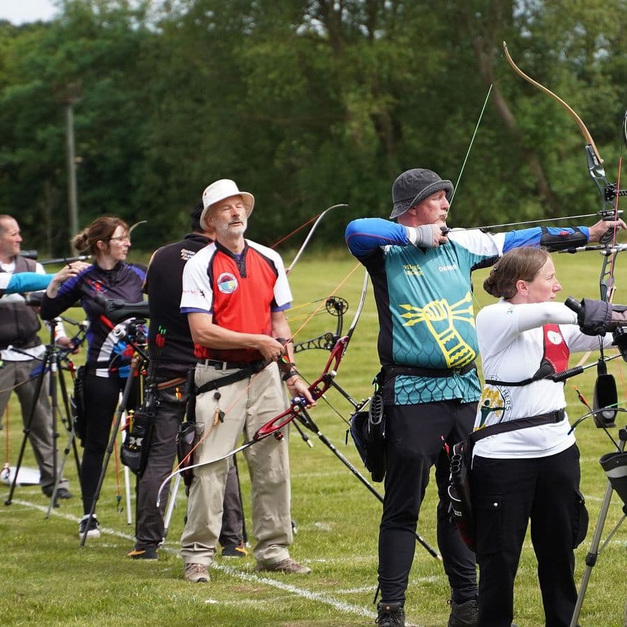 A line of archers at full draw shooting recurve and compound bows across a grass field, with trees in the background.