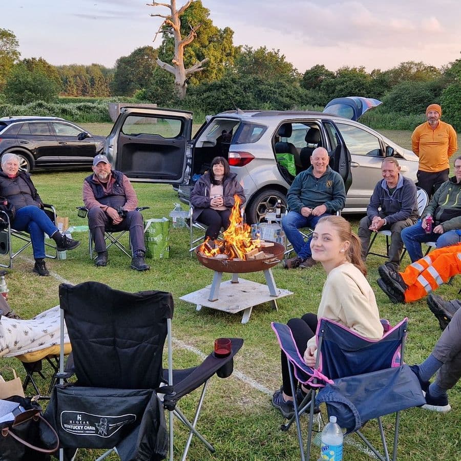 A group of around twenty people sitting in camping chairs around a lit fire pit on a grass field at dusk, socialising with drinks, with cars parked behind them.