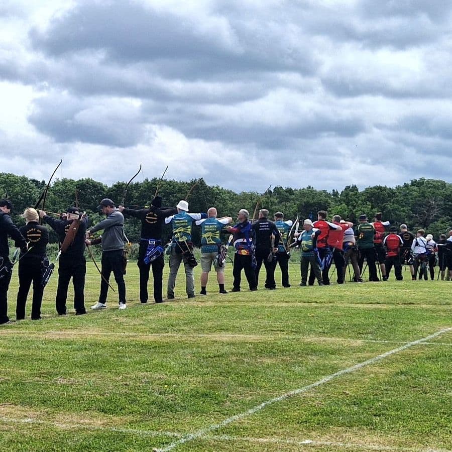 A very wide view of dozens of archers at full draw in a long shooting line under a cloudy sky, with targets and trees in the distance.