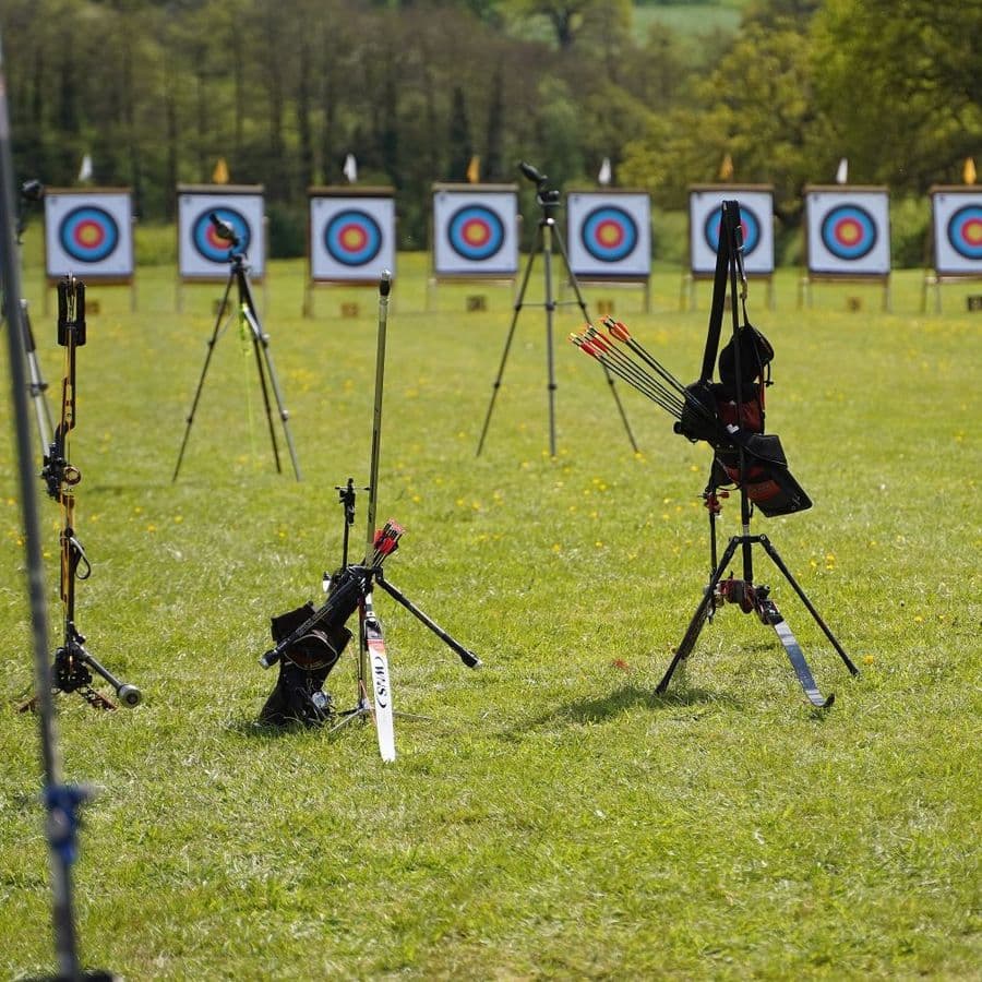 Compound and recurve bows resting on stands with quivers of arrows, in front of a long row of numbered archery targets.