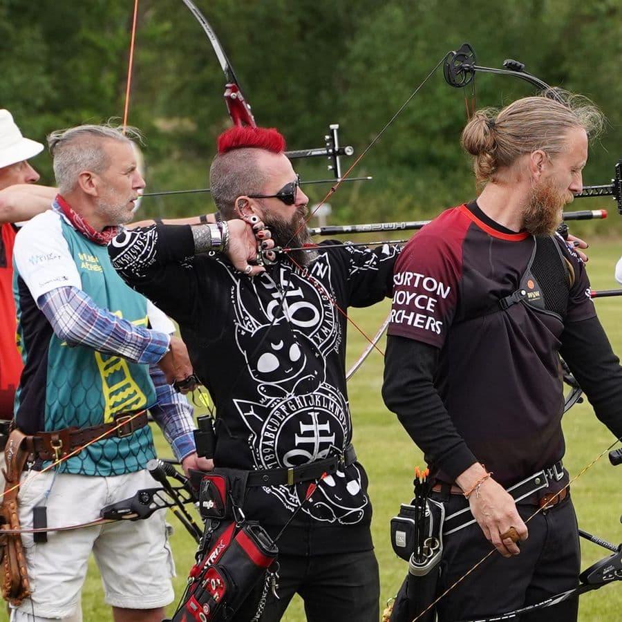 A row of archers shooting bows, including one with a bright red mohawk and tattoos wearing a Burton Joyce Archers shirt at full draw.