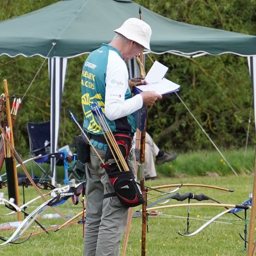 An archer in a Welbeck Archers shirt checking a scoresheet beside a row of recurve bows.