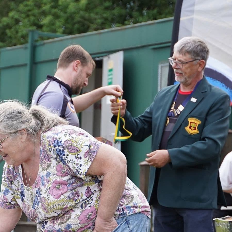 A club official in a green blazer with medals handing out a green and yellow ribboned award, with an older woman and others nearby, trophies visible on a table.