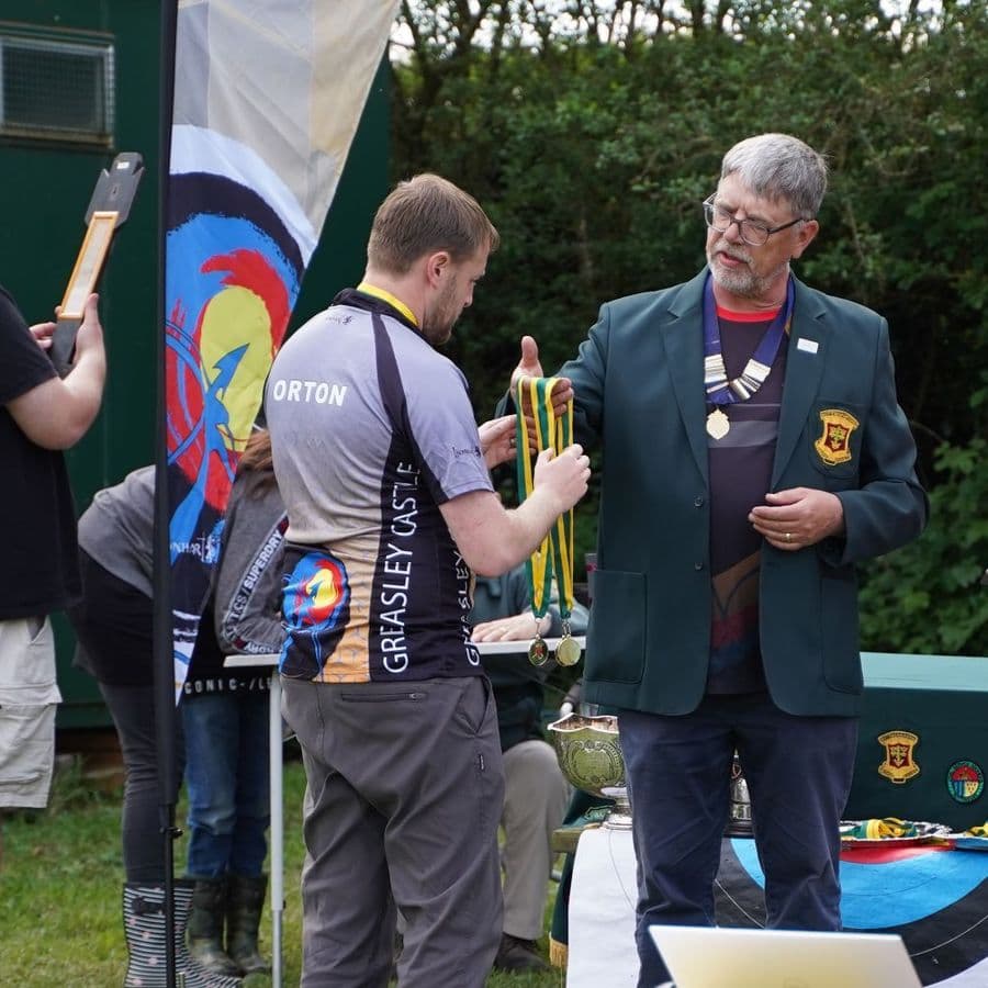 A club official presenting medals to an archer at an outdoor awards presentation.