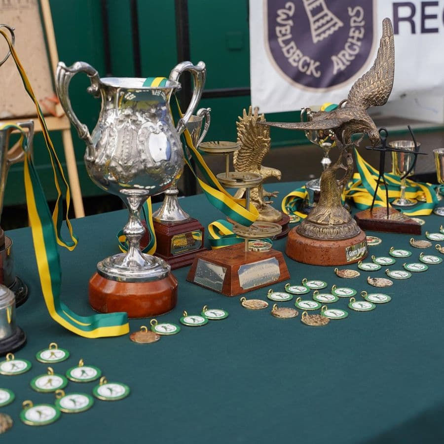 A table displaying archery trophies, cups and medals with green and yellow ribbons, in front of a Welbeck Archers banner.