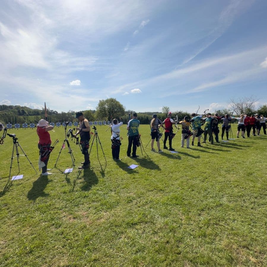 A wide view of a long line of archers at full draw across a grass field under a bright sky, targets in the distance.