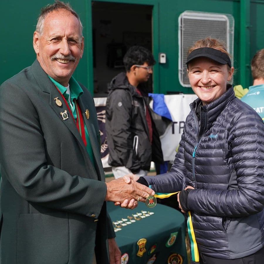 A club official in a green blazer shaking hands with an archer and presenting her with a medal on a green and yellow ribbon.