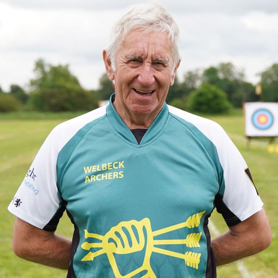 A smiling Welbeck Archers member in a team shirt standing on the range with a target visible behind him.