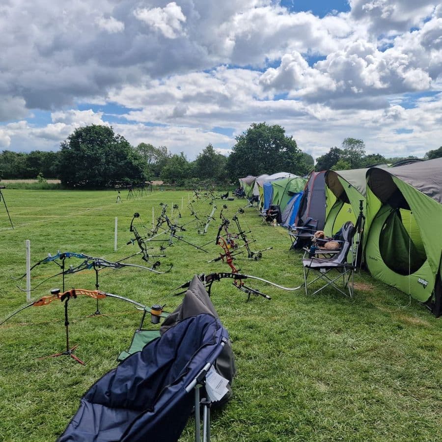 A long row of recurve and compound bows resting on stands in front of a line of camping tents pitched on a grass field, with spotting scopes set up on the archery range in the background.