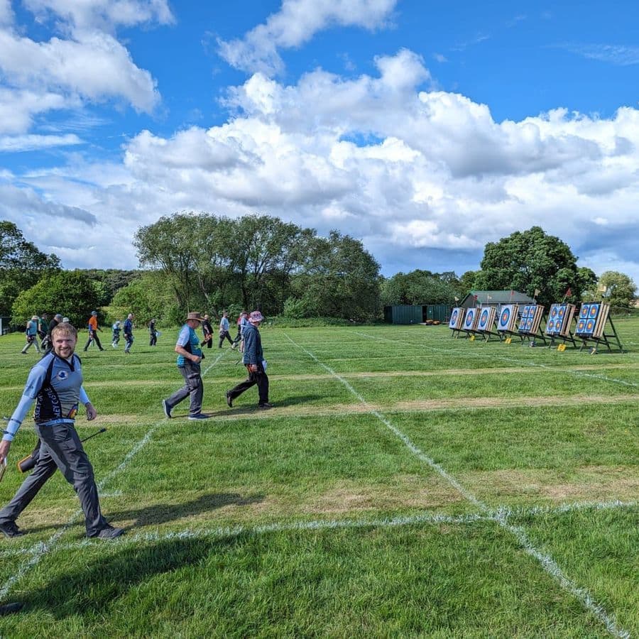 Archers walking across a grass field carrying scorecards, with a row of archery targets set up in the distance under a partly cloudy sky.