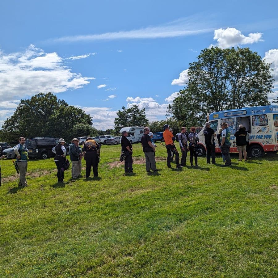 A group of archers queuing at an ice cream van parked on the grass field, with camper vans and cars parked behind them.
