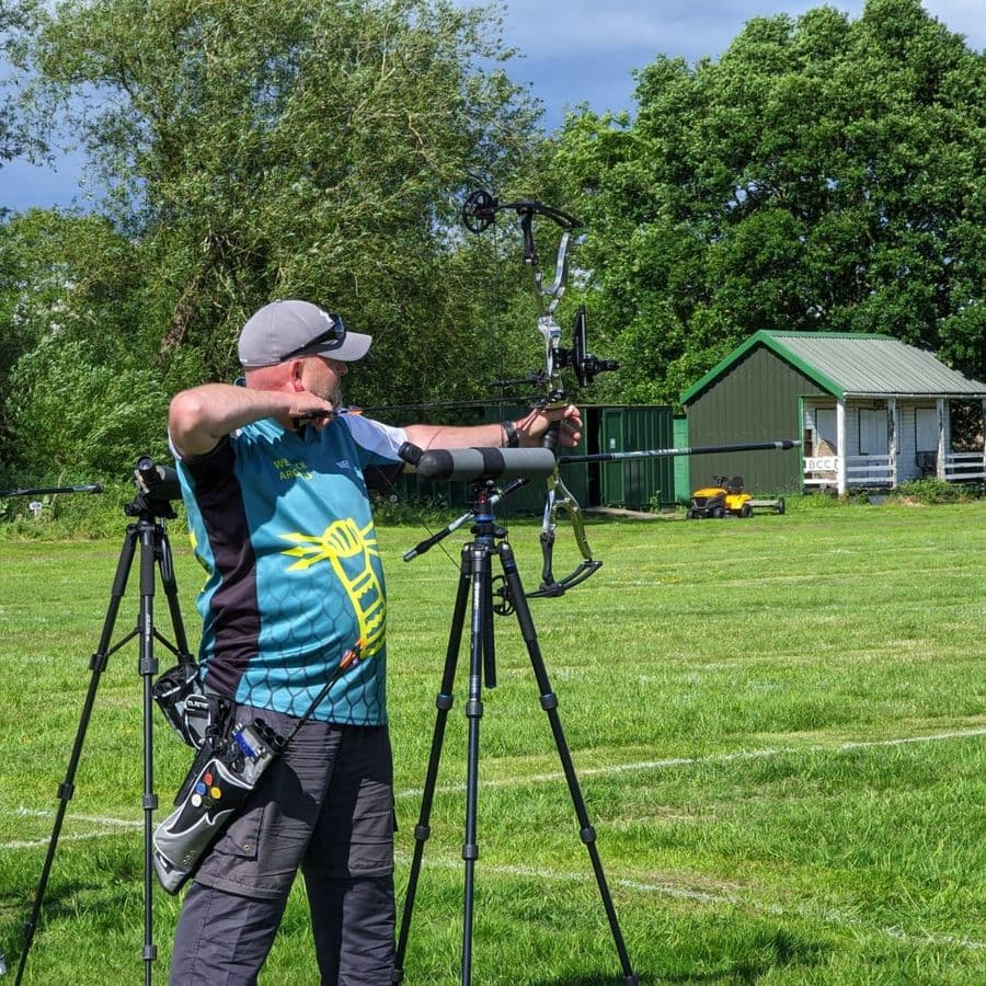 A man in a Welbeck Archers jersey at full draw with a compound bow, aiming down the field past camera tripods, with a green-roofed shed in the background.