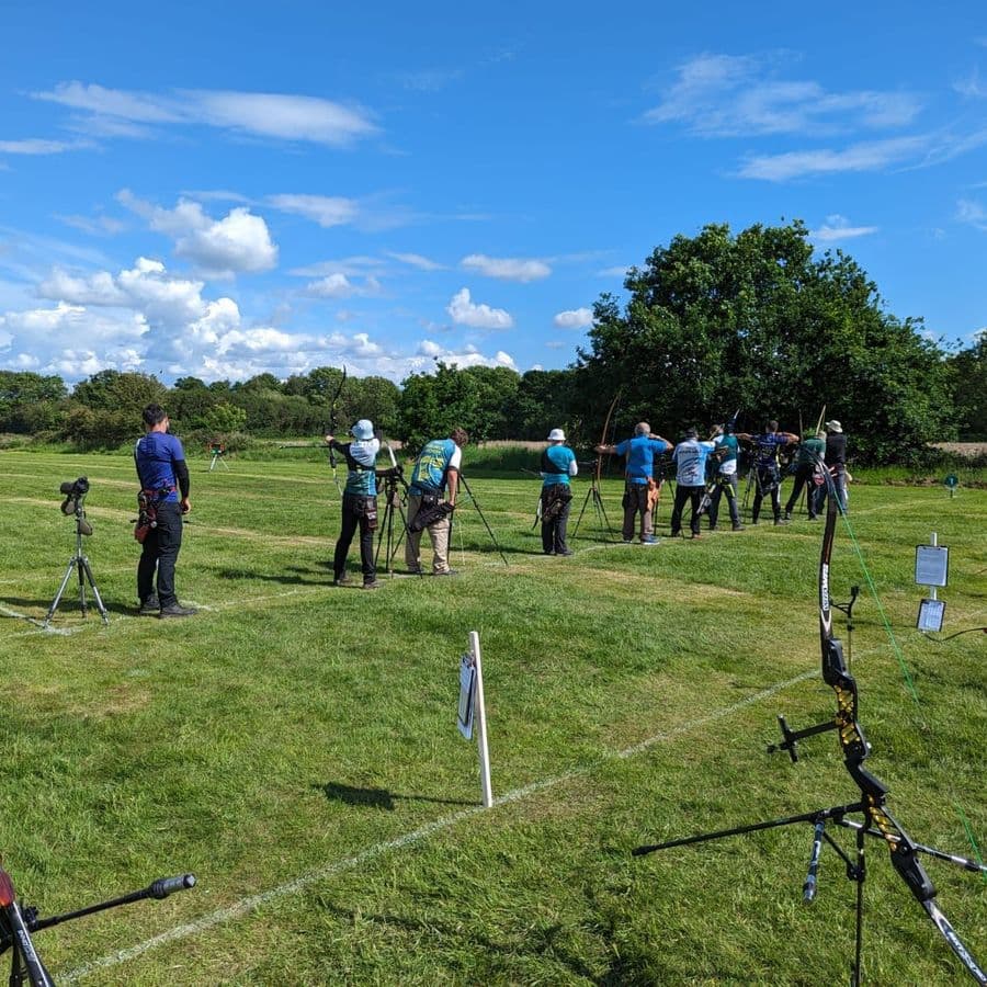 A line of archers shooting recurve bows at targets on a sunny day, with scorecards on stands and camera tripods set up along the shooting line.
