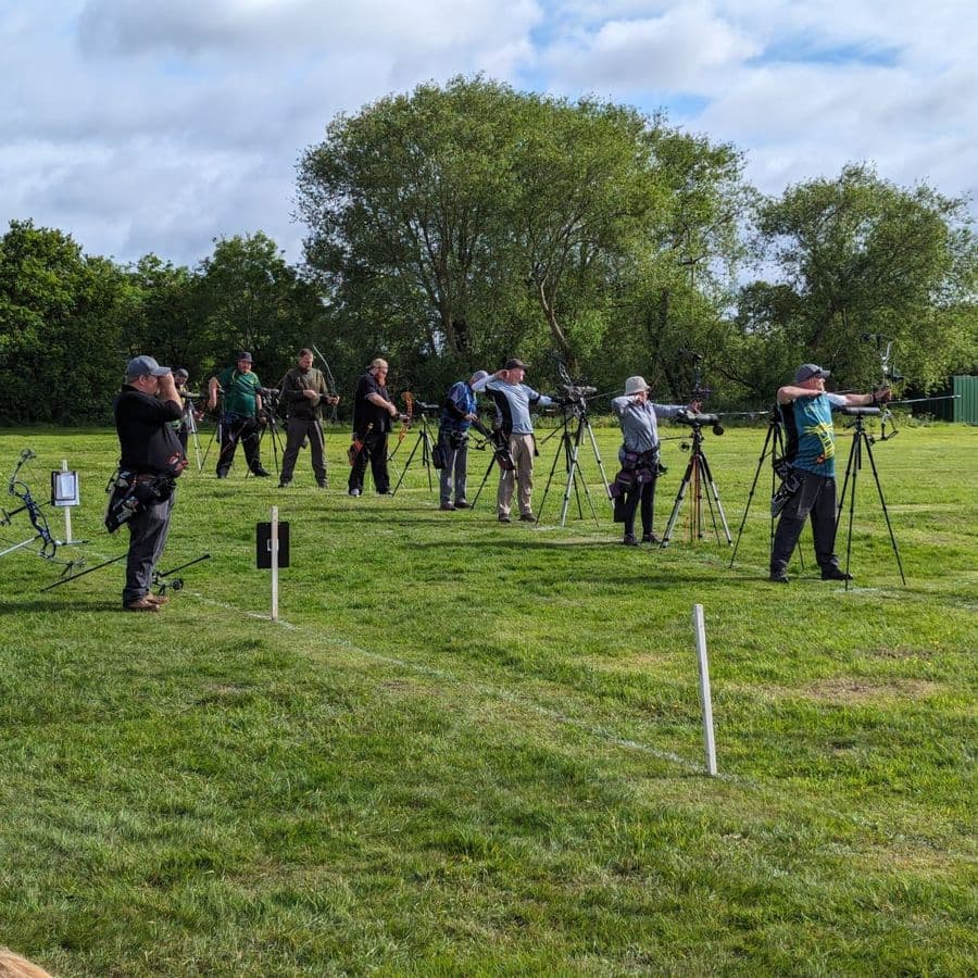 A line of about a dozen archers shooting compound and recurve bows on a grass field, with camera tripods and a scoreboard set up along the line.