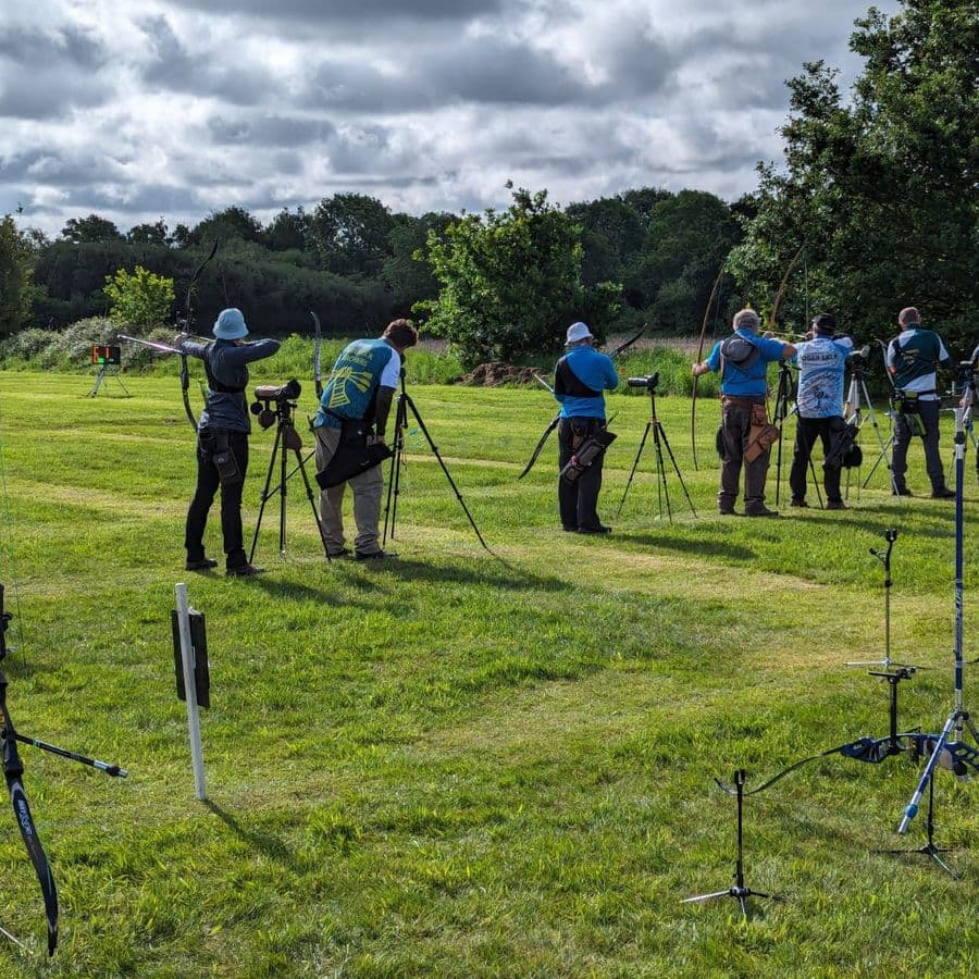 A row of archers at full draw shooting recurve bows across a field, with spotting scopes on tripods and trees in the background.