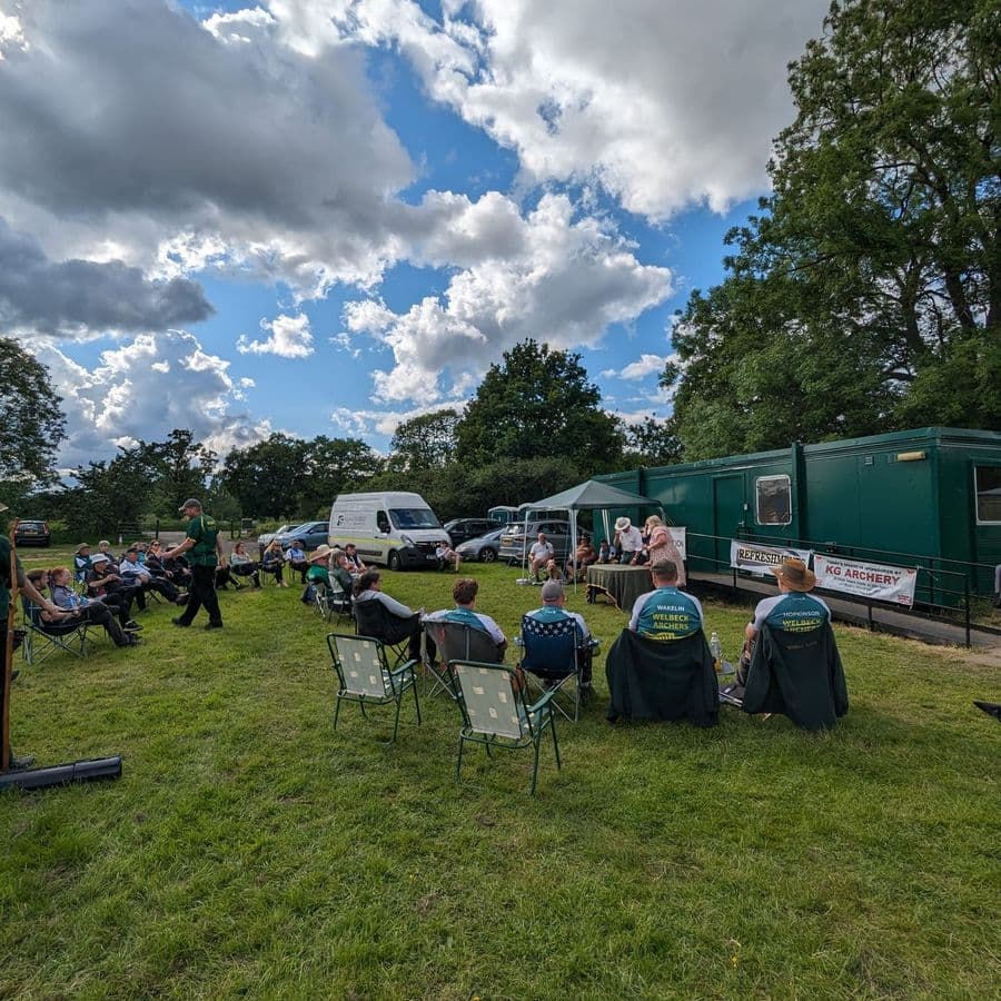 A crowd of archers sitting in camping chairs on the grass listening to an announcement near a green portable building with a "Refreshments" banner and a KG Archery sponsor sign, campervans parked nearby.