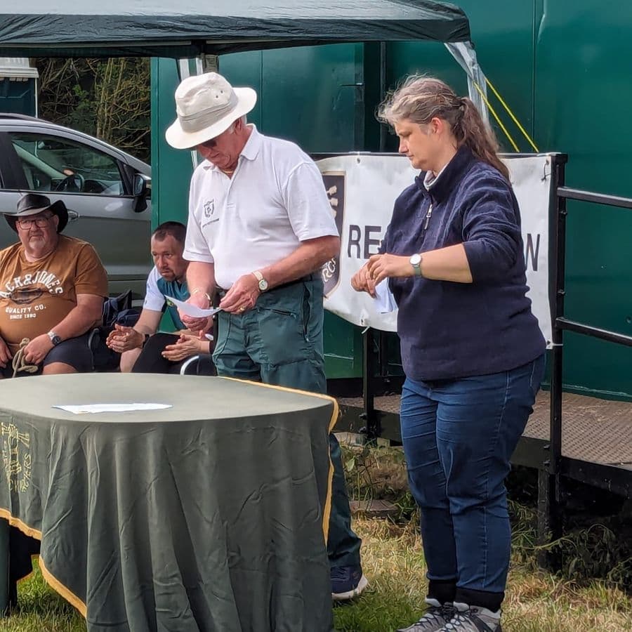 A man in a wide-brimmed hat and a woman standing at a table reading out results, with seated archers and camper vans in the background.