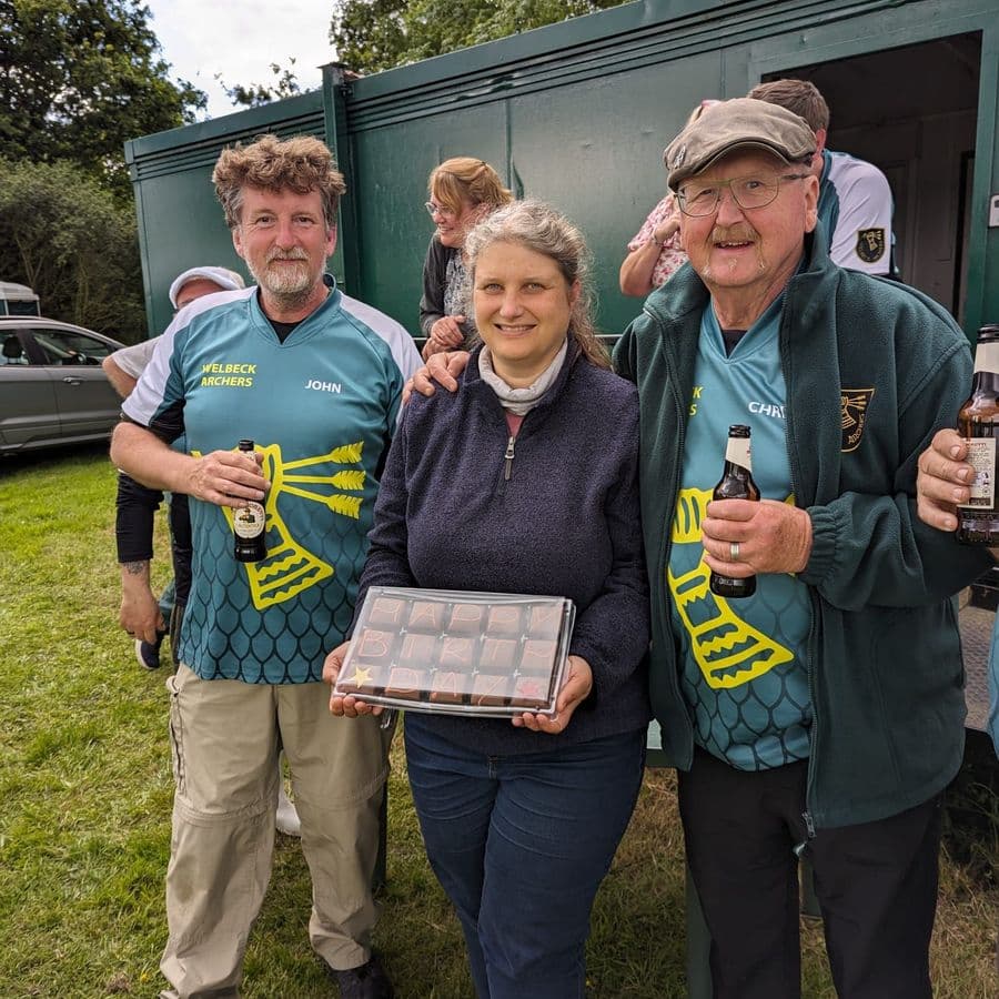 Three Welbeck Archers members in club jerseys smiling for a photo, holding beer bottles and a chocolate box decorated "Happy Birthday", in front of a green cabin.