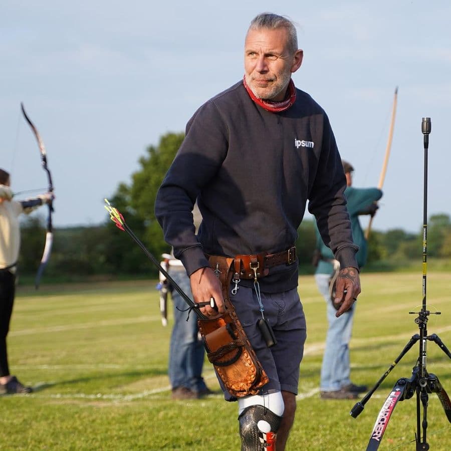 A man with a prosthetic lower leg stands on the archery field holding arrows, with other archers shooting recurve bows and a target visible in the background.