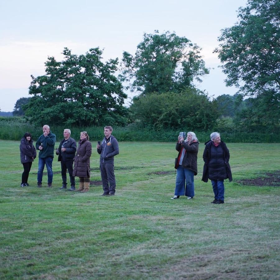 A group of people standing on a grass field at dusk, looking upward, some holding up phones to take photos.