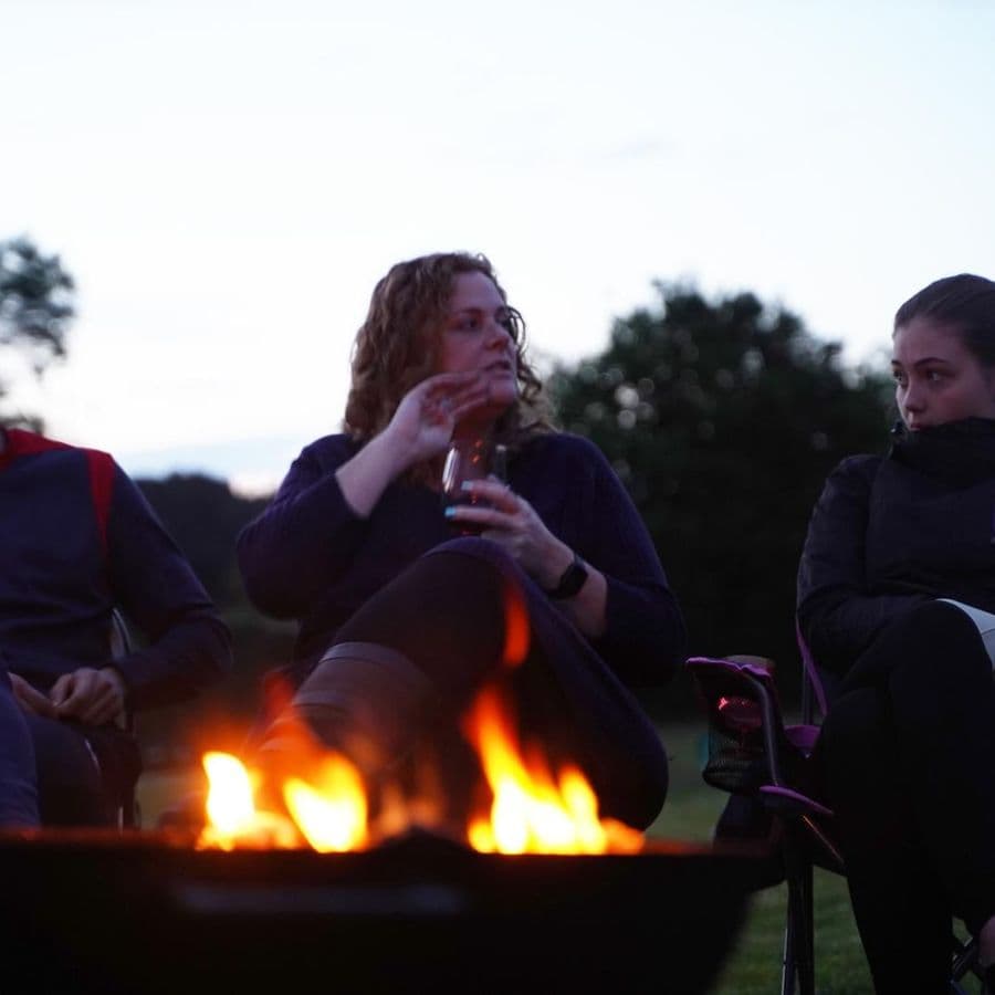 Three people sitting around a lit fire pit at dusk, one holding a drink.