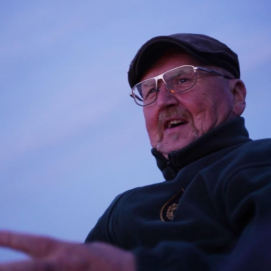 Close-up portrait of an older man wearing glasses and a flat cap, smiling and gesturing, photographed at dusk.
