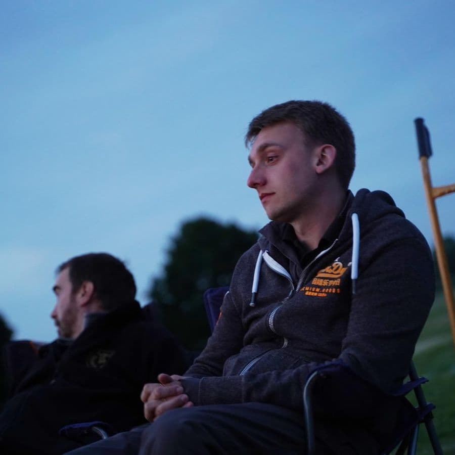 Two young men sitting in camping chairs on a field at dusk, one wearing a dark hoodie.