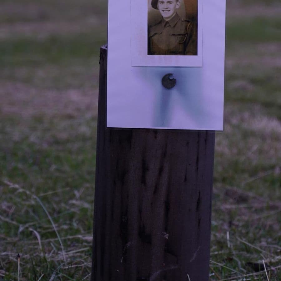 A black-and-white portrait photo of a young soldier in WWII uniform, mounted on a wooden post in a field as a memorial display, with a poppy pinned beneath it.