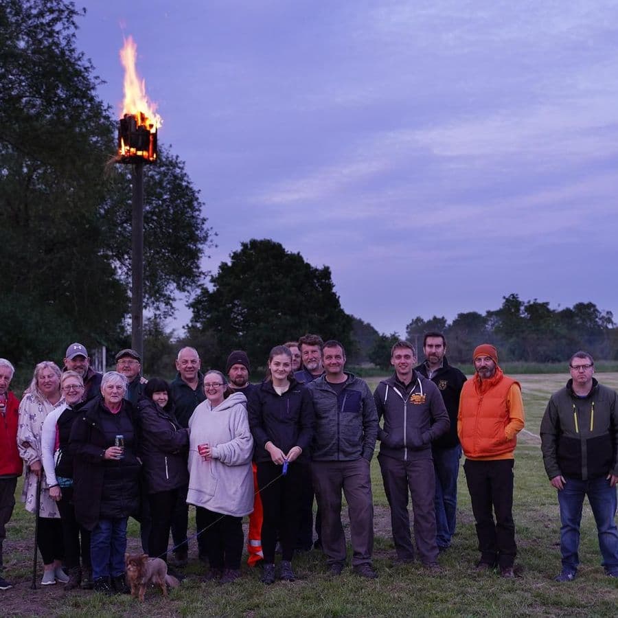 A group of around fifteen people standing on a grass field at dusk beneath a tall pole topped with a lit beacon brazier, part of the D-Day 80th anniversary commemoration.