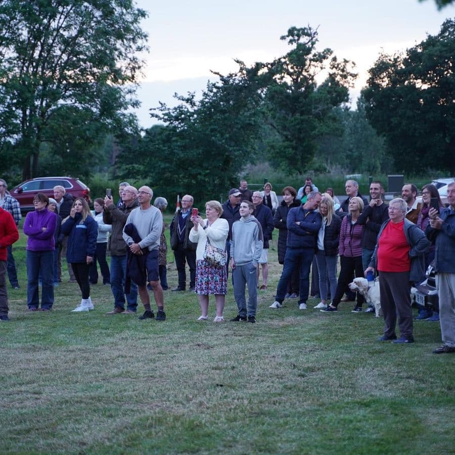 A crowd of people standing on a grass field at dusk near parked cars, looking upward, some holding up phones.