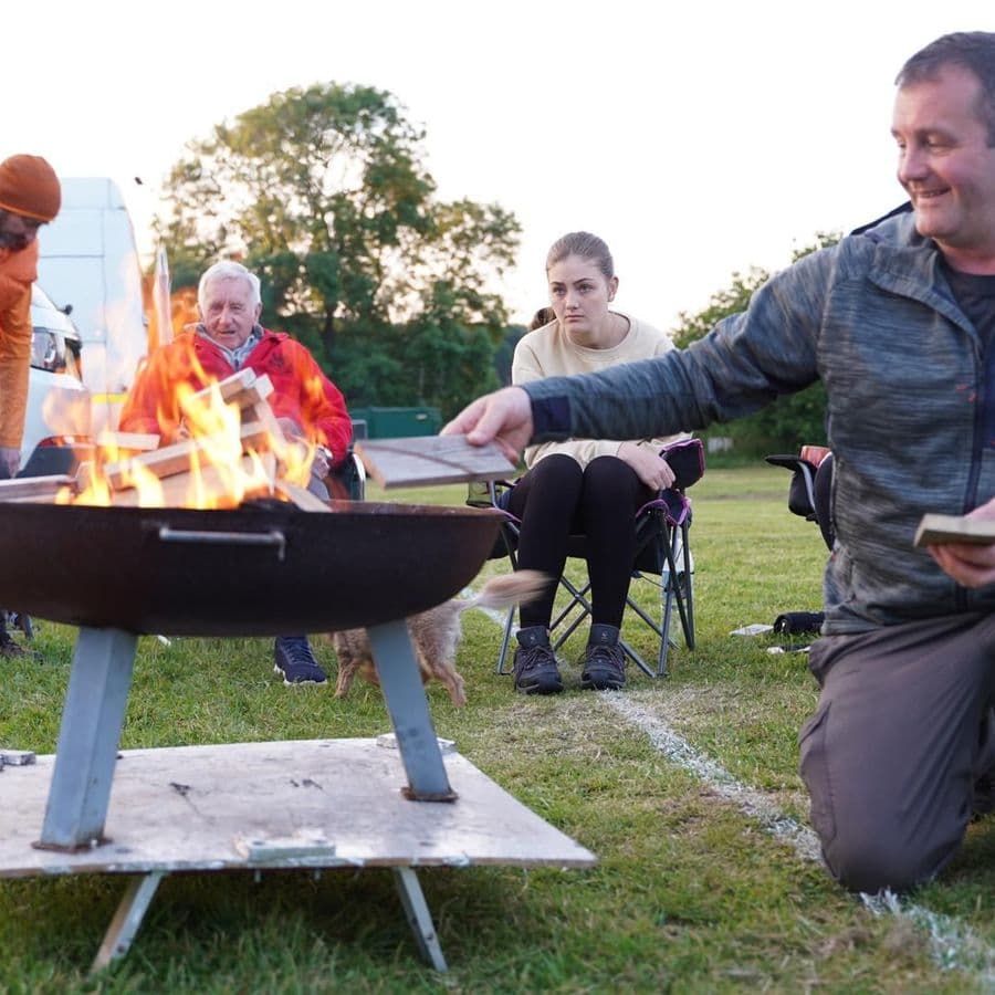 A group of people gathered around a lit fire pit on a grass field at dusk, one man kneeling and smiling while adding wood to the fire, a dog nearby.