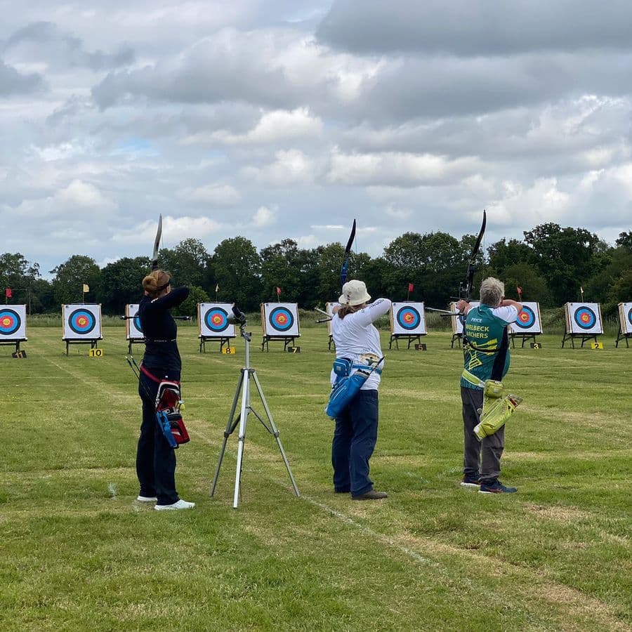 Three archers standing at the shooting line taking aim with recurve bows, with a numbered row of archery targets in front of them, one wearing a Welbeck Archers jersey.