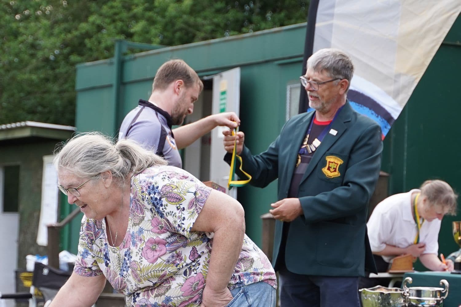 A club official in a green blazer with medals handing out a green and yellow ribboned award, with an older woman and others nearby, trophies visible on a table.