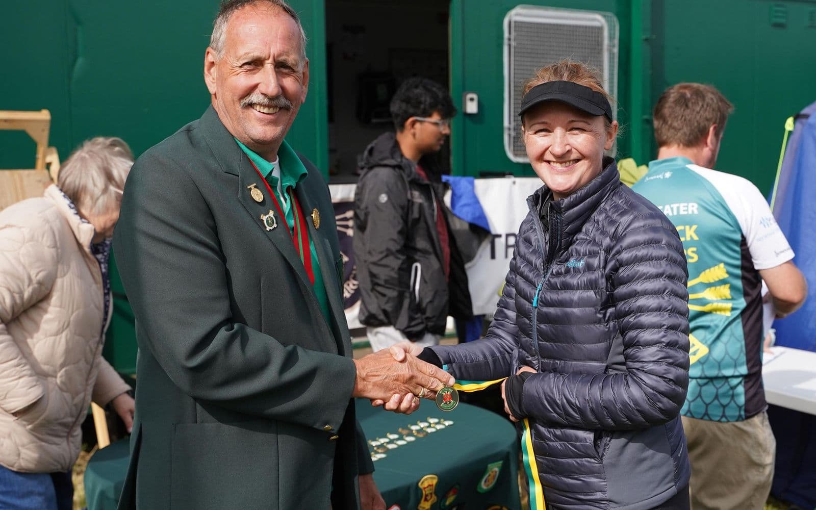 A club official in a green blazer shaking hands with an archer and presenting her with a medal on a green and yellow ribbon.