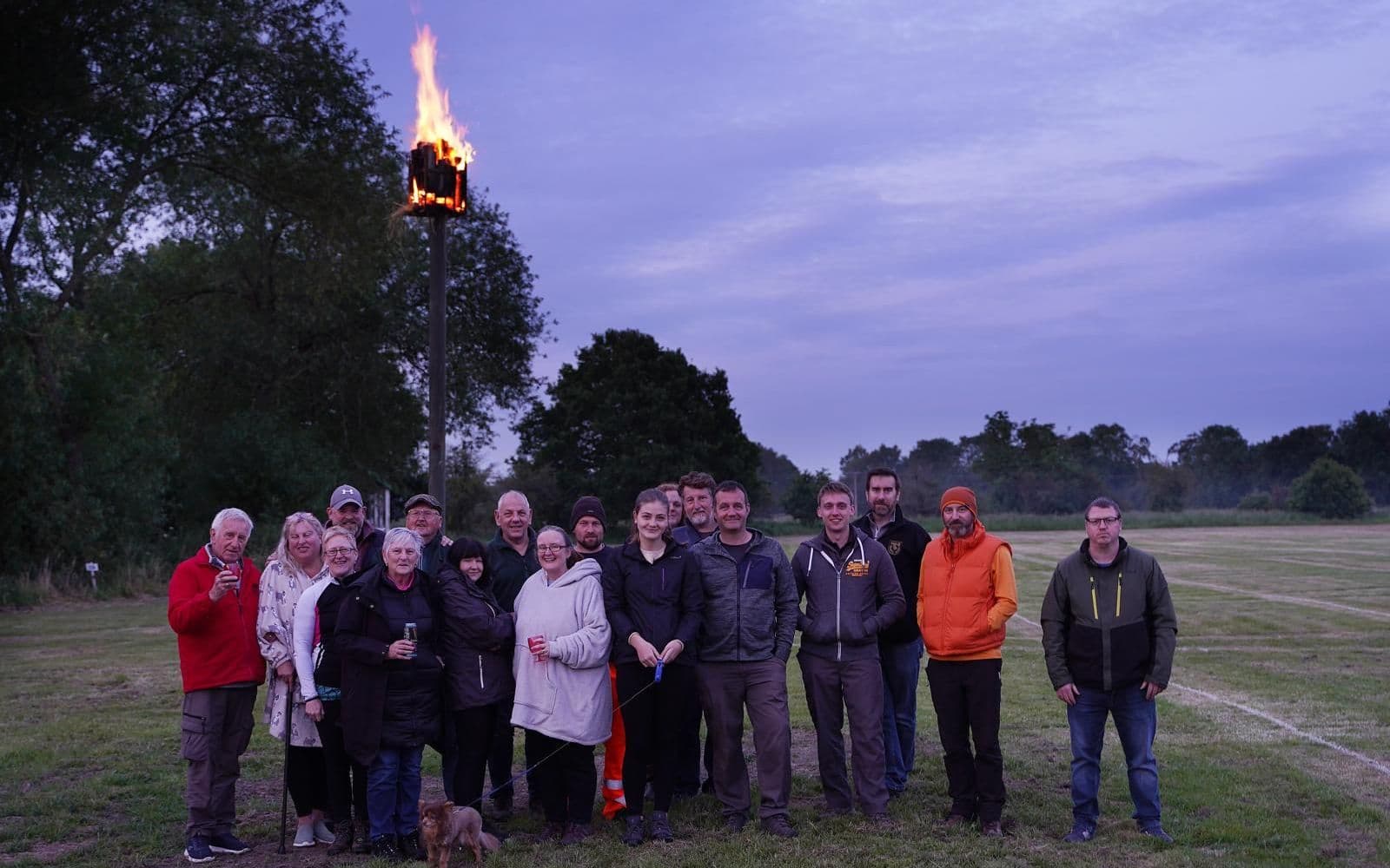 A group of around fifteen people standing on a grass field at dusk beneath a tall pole topped with a lit beacon brazier, part of the D-Day 80th anniversary commemoration.